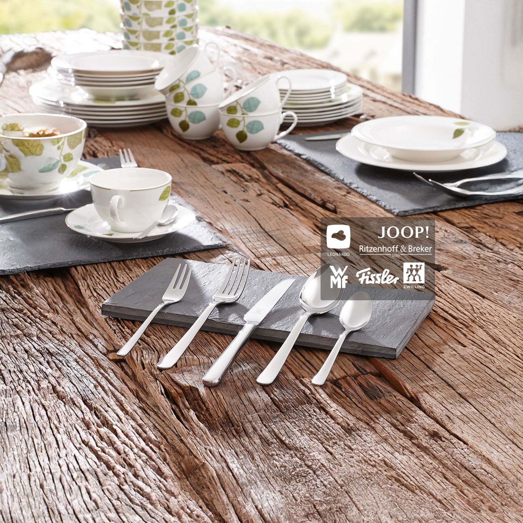 Gedeckter Holztisch mit weißem Geschirr mit Blattmotiv und glänzendem Besteck auf Schieferplatten im natürlichen Ambiente