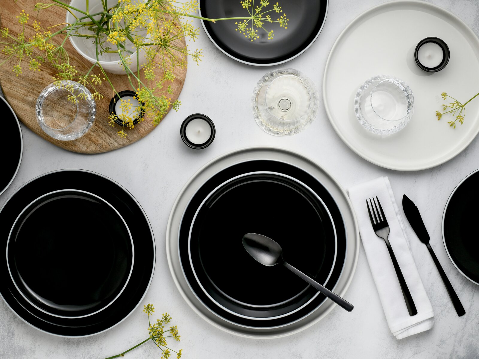 Modernes Tischgedeck mit schwarzen Tellern, Glas-Schalen, schwarzem Besteck und kleinen Kerzen auf heller Oberfläche, dekoriert mit gelben Blüten – Draufsicht (Flatlay)