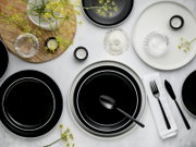 Modernes Tischgedeck mit schwarzen Tellern, Glas-Schalen, schwarzem Besteck und kleinen Kerzen auf heller Oberfläche, dekoriert mit gelben Blüten – Draufsicht (Flatlay)