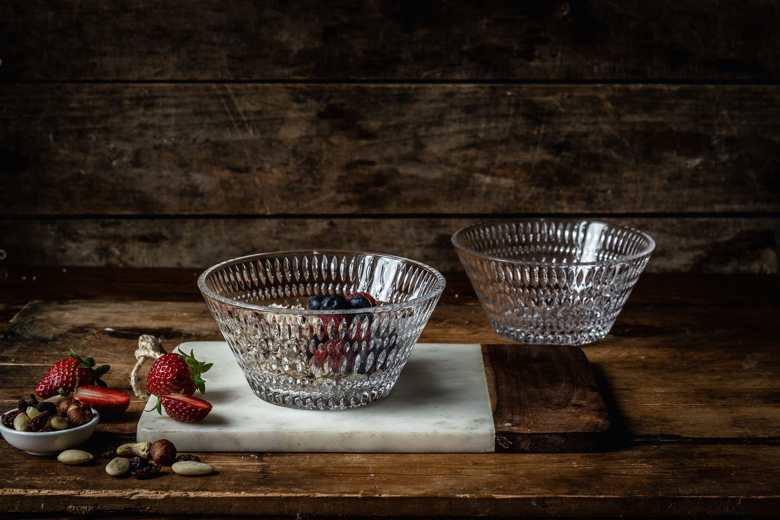 Zwei durchsichtige Glasschalen mit Ethno-Muster, eine gefüllt mit Beeren und Nüssen, auf einem rustikalen Holztisch von schräg oben fotografiert.