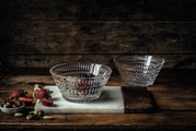 Twee doorzichtige glazen schalen met een etnisch patroon, waarvan er één gevuld is met bessen en noten, gefotografeerd vanaf een schuine hoek bovenop een rustieke houten tafel.