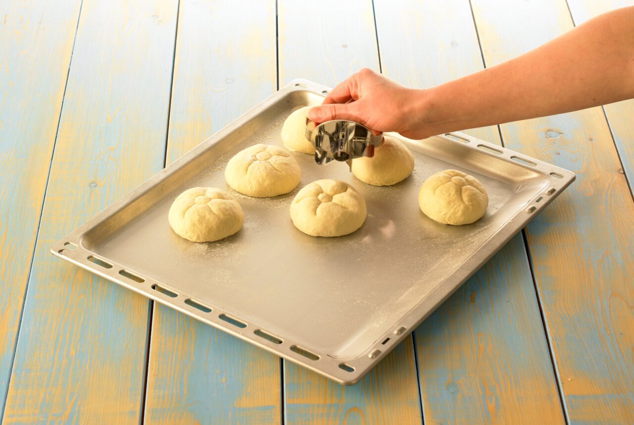 Een hand stempelt broodjes op een bakplaat met een broodjestempel, gefotografeerd van bovenaf.