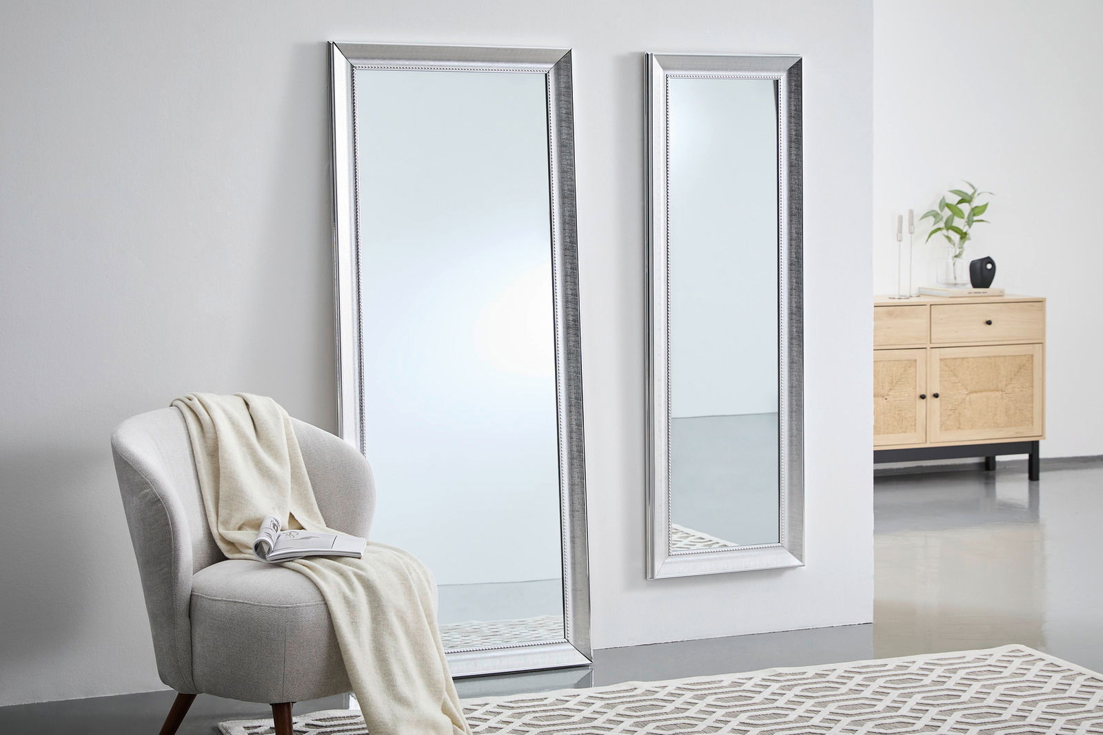 Zwei große, rechteckige Spiegel mit silbernem Rahmen, einer steht auf dem Boden, der andere ist an der Wand montiert. Perspektive von schräg vorne.