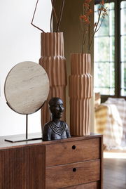 Zwei hohe, geriffelte Vasen in Erdtönen mit Zweigen und Blumen, daneben eine Büste und ein runder, steinartiger Dekorationsgegenstand auf einem Holzschrank. Perspektive: seitlich.