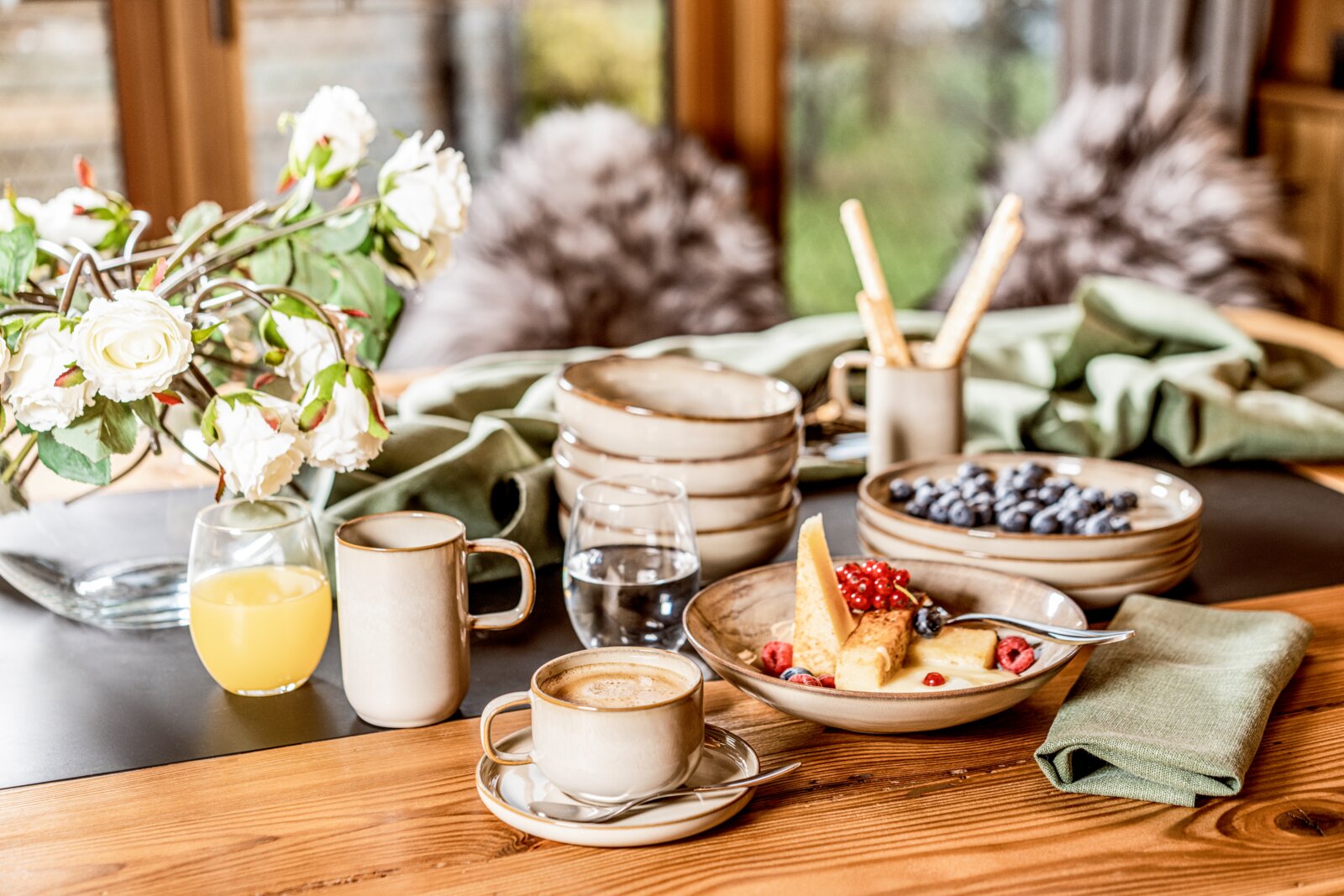 Gipfelstück Dessertteller 4er-Set NEBELWALD Gemütlich gedeckter Frühstückstisch mit beigefarbenem Keramikgeschirr, Kaffee, Saft, Wasser, Blaubeeren und Dessertschale neben Blumen und grüner Stoffserviette, aufgenommen aus leicht erhöhter schräger Vorderansicht.