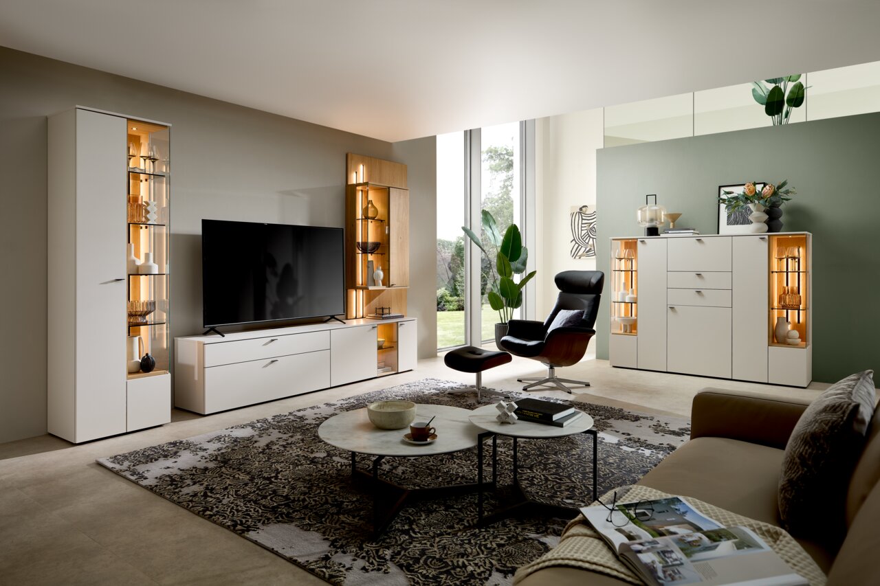 Moderne Wohnzimmeransicht mit eleganter Vitrine in hellem Holz und Glas, links im Bild, mit dekorativer Beleuchtung und stilvollen Accessoires.