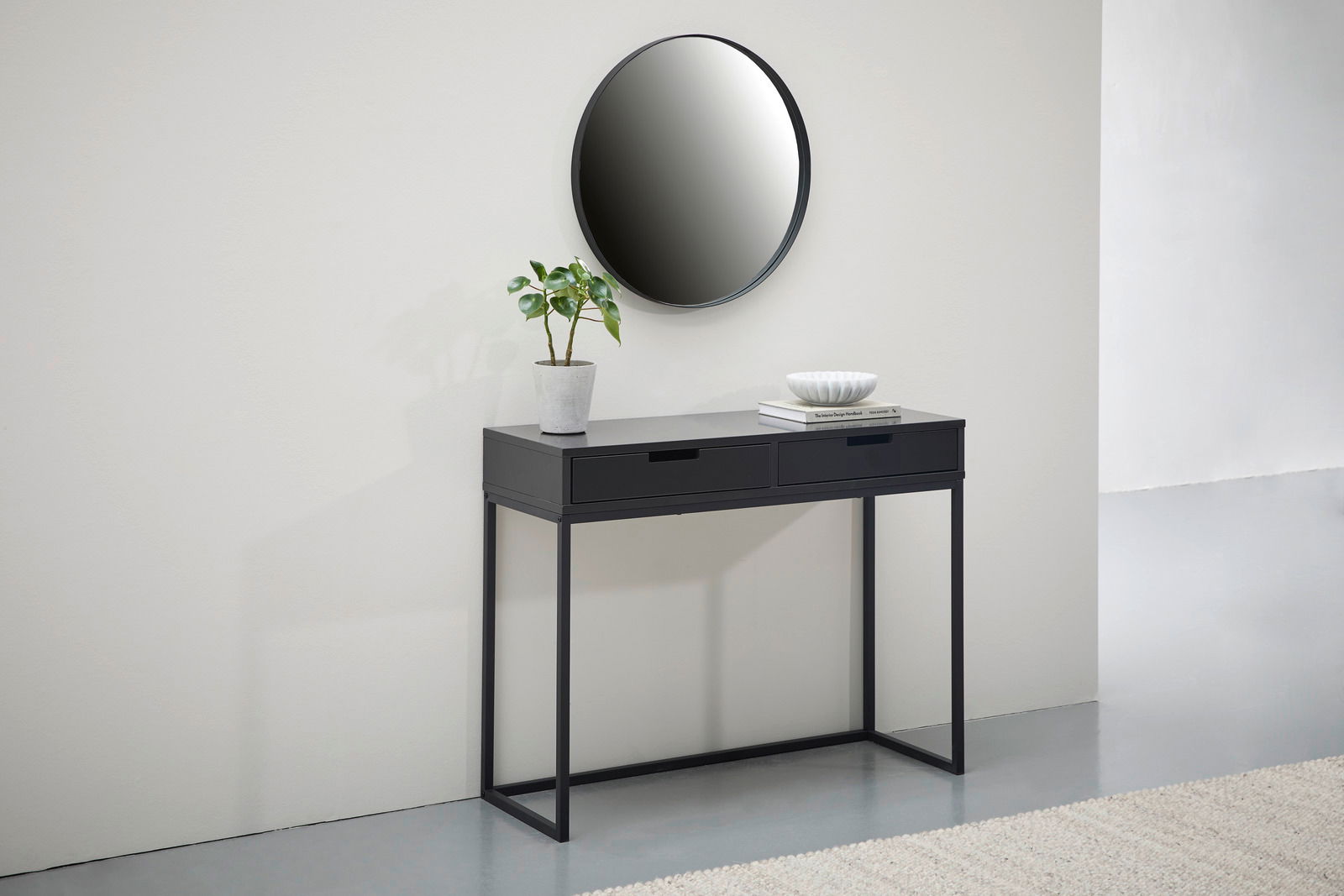 Schwarze Konsole mit zwei Schubladen, dekoriert mit einer Pflanze und einer Schale, vor einem runden Spiegel an der Wand. Frontalansicht.