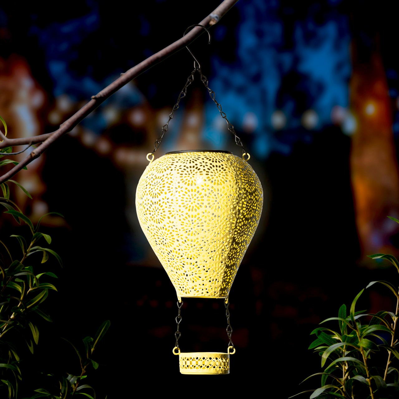 Metallener Solarballon in Form eines Heißluftballons, aufgehängt an einem Ast, leuchtet in der Dämmerung. Perspektive von vorne.