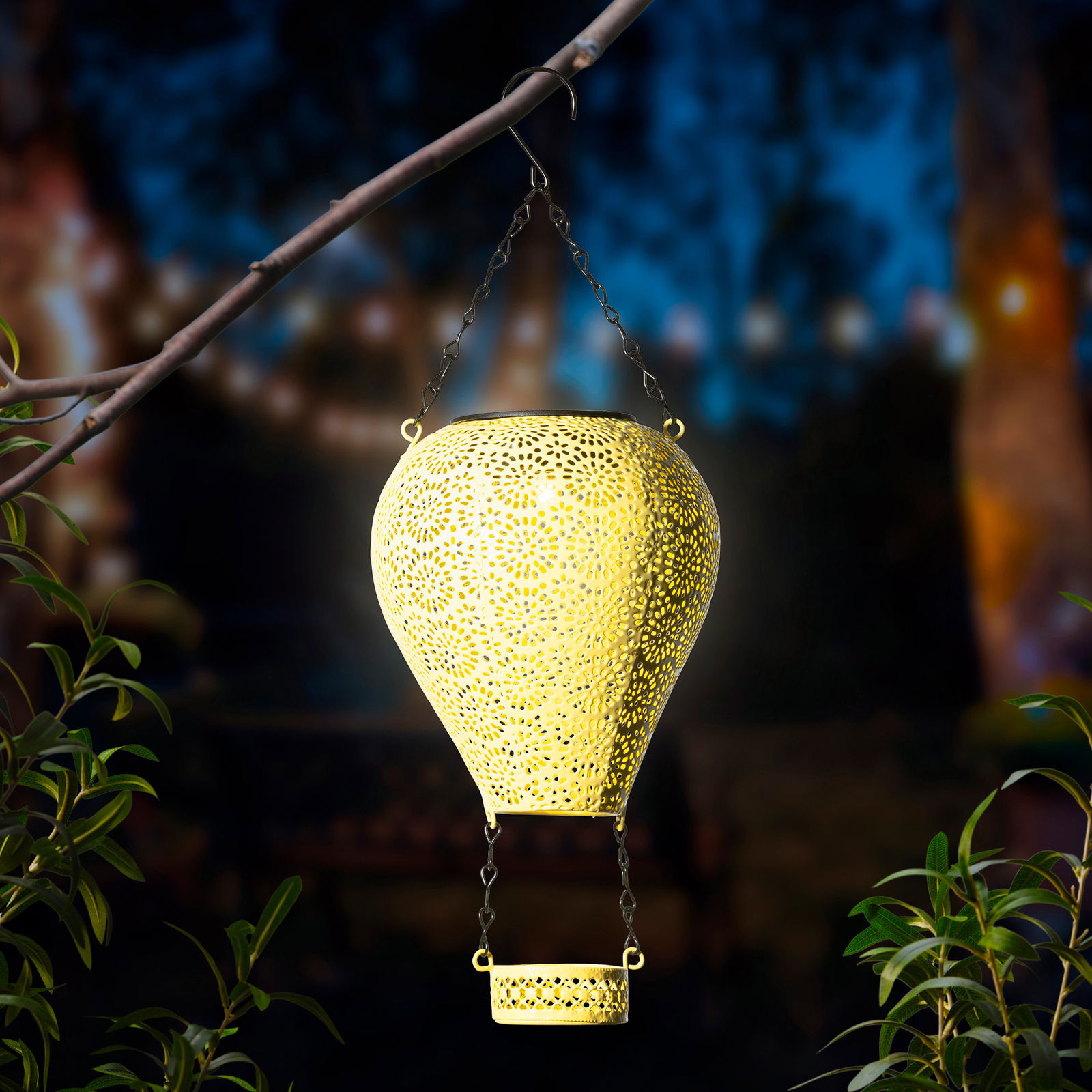 Metallener Solarballon in Form eines Heißluftballons, aufgehängt an einem Ast, leuchtet in der Dämmerung. Perspektive von vorne.