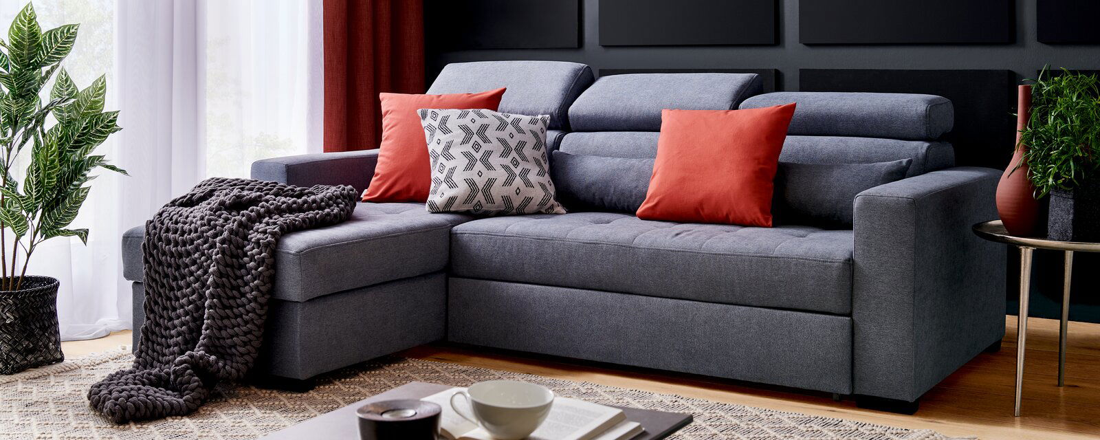 Graue Rundecke mit roten und gemusterten Kissen, dekoriert mit einer gestrickten Decke, seitliche Perspektive