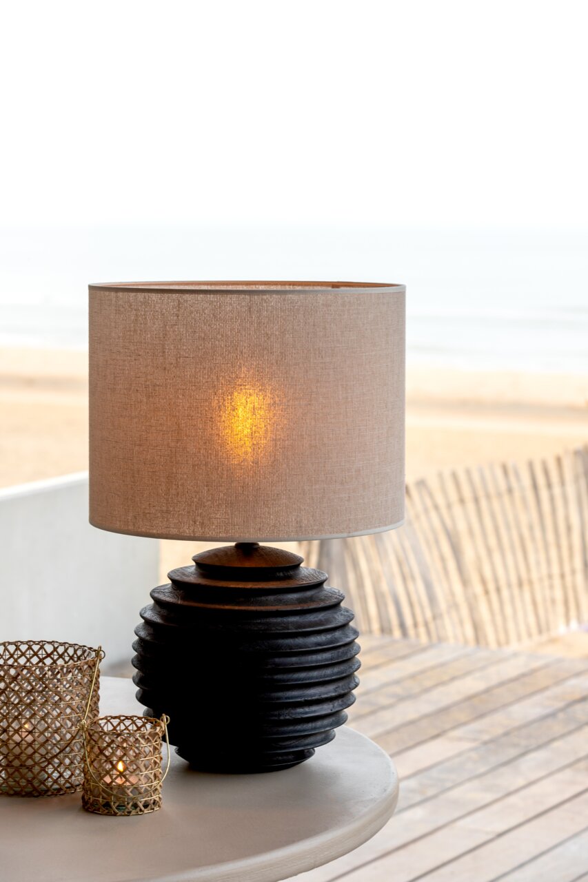 Moderner Lampenschirm auf einem gerippten, dunklen Sockel, seitliche Perspektive mit Strandhintergrund.