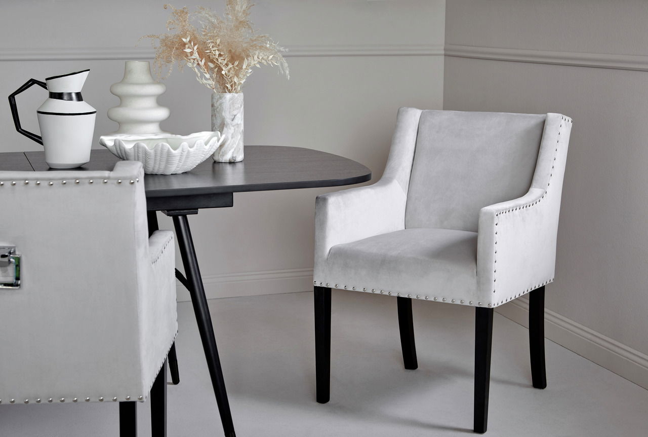 Eleganter grauer Stuhl mit schwarzen Beinen und Nietenverzierung, seitliche Perspektive, neben einem runden Tisch mit dekorativen Vasen.