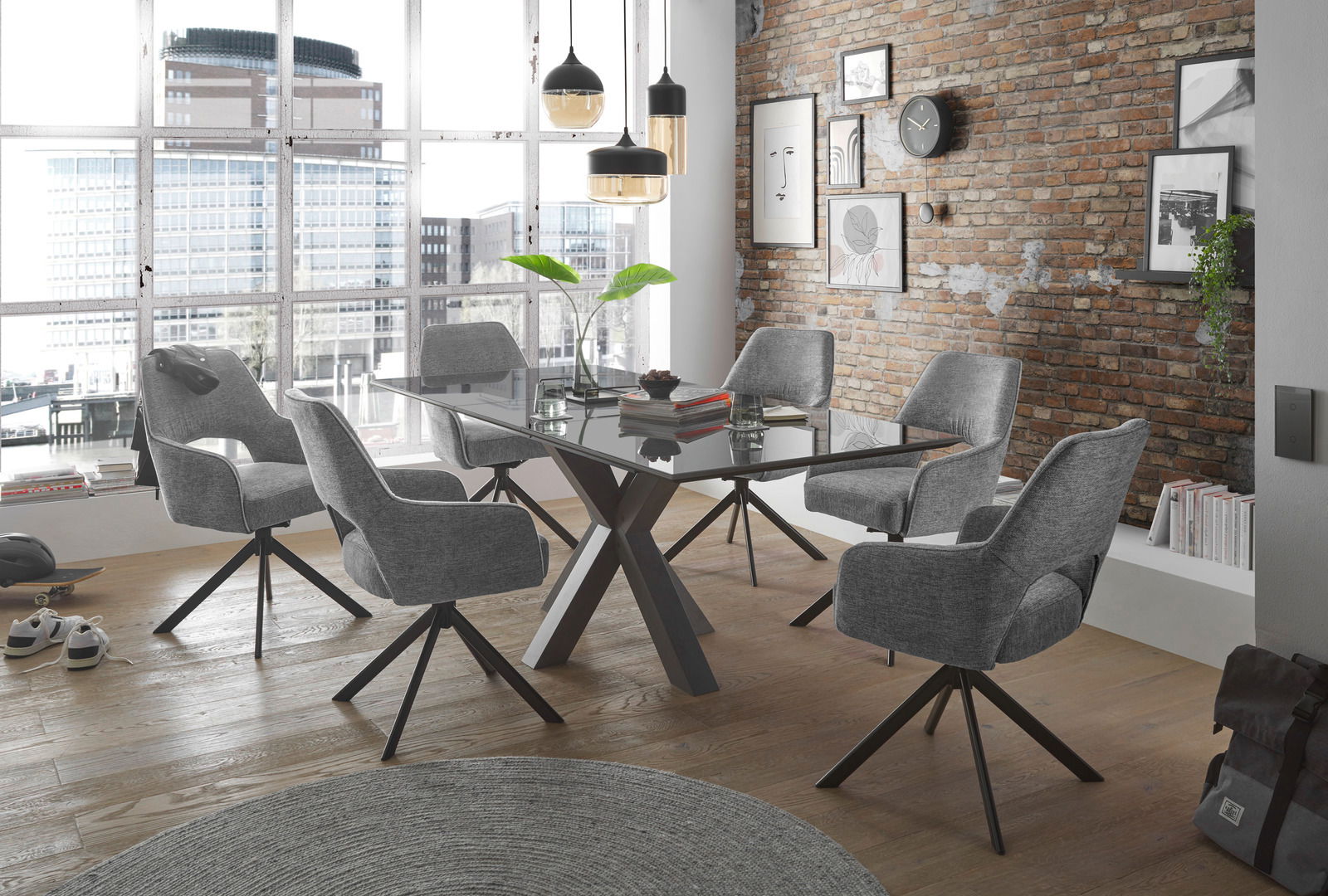 Set one by Musterring Armlehnstuhl SET ONE WINSTON Moderner Essbereich mit sechs grauen Armlehnstühlen um einen Glastisch, aus der Perspektive eines offenen, hellen Raumes mit Backsteinwand und großen Fenstern.