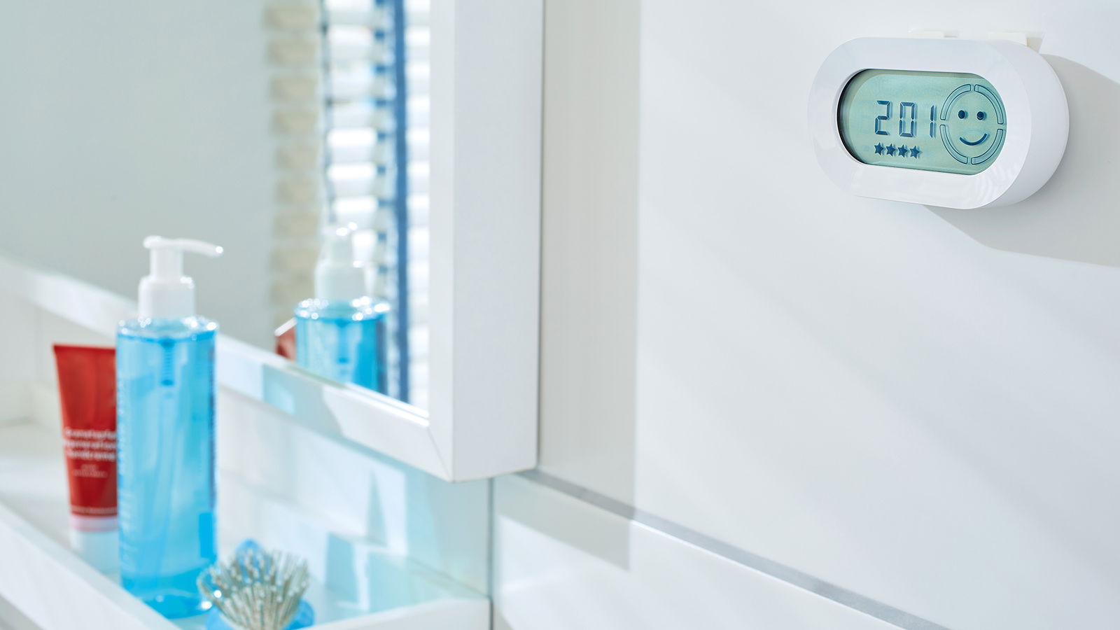 Badezimmeransicht mit einem digitalen Thermometer an der Wand, das 20,1 Grad anzeigt, und einem lächelnden Gesichtssymbol. Im Vordergrund sind ein blauer Seifenspender und ein rotes Produkt zu sehen.