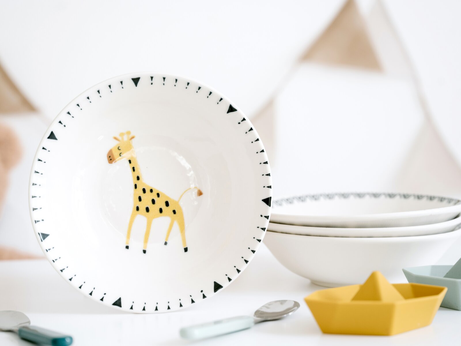 Weiße Kinderschale aus Keramik mit Giraffen-Motiv und schwarzem Dreieckmuster am Rand, frontal in leichter Schrägansicht fotografiert; im Hintergrund gestapelte Schalen und kleines gelbes Origami-Boot.