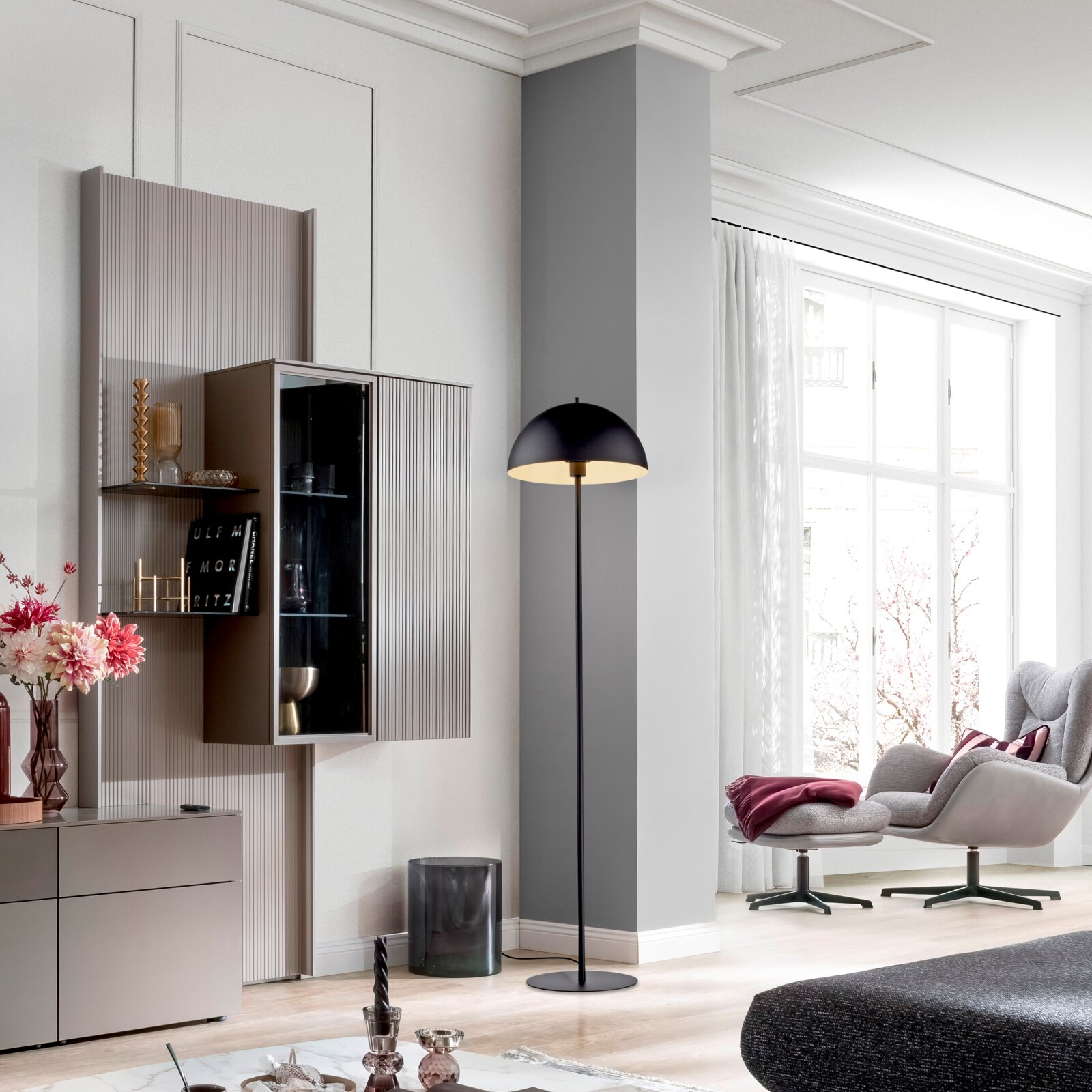 Moderne schwarze Stehleuchte mit halbrundem Schirm und warmem Licht in einem hellen Wohnzimmer neben einem Sessel, frontale Perspektive.