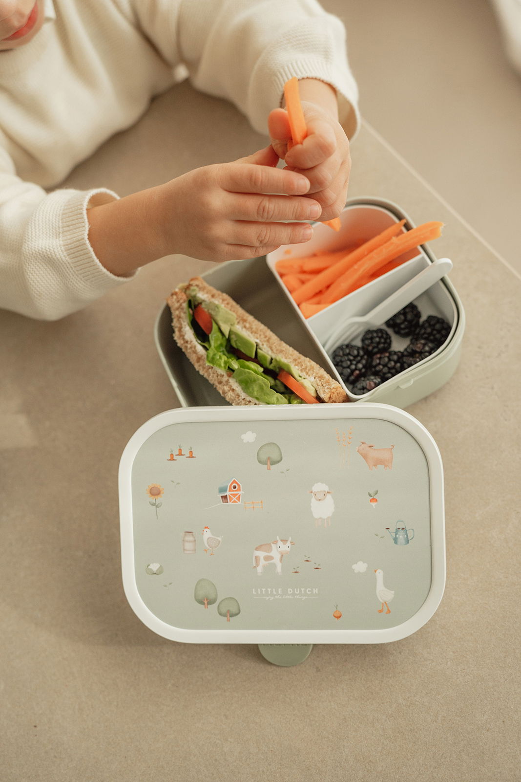 Lunchset Campus Little Farm mit kindgerechtem Design, bestehend aus einer Brotdose mit Bauernhofmotiven und einem Kind, das Karottensticks hält. Perspektive von oben.