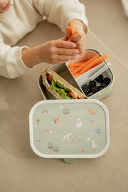 Lunchset Campus Little Farm mit kindgerechtem Design, bestehend aus einer Brotdose mit Bauernhofmotiven und einem Kind, das Karottensticks hält. Perspektive von oben.