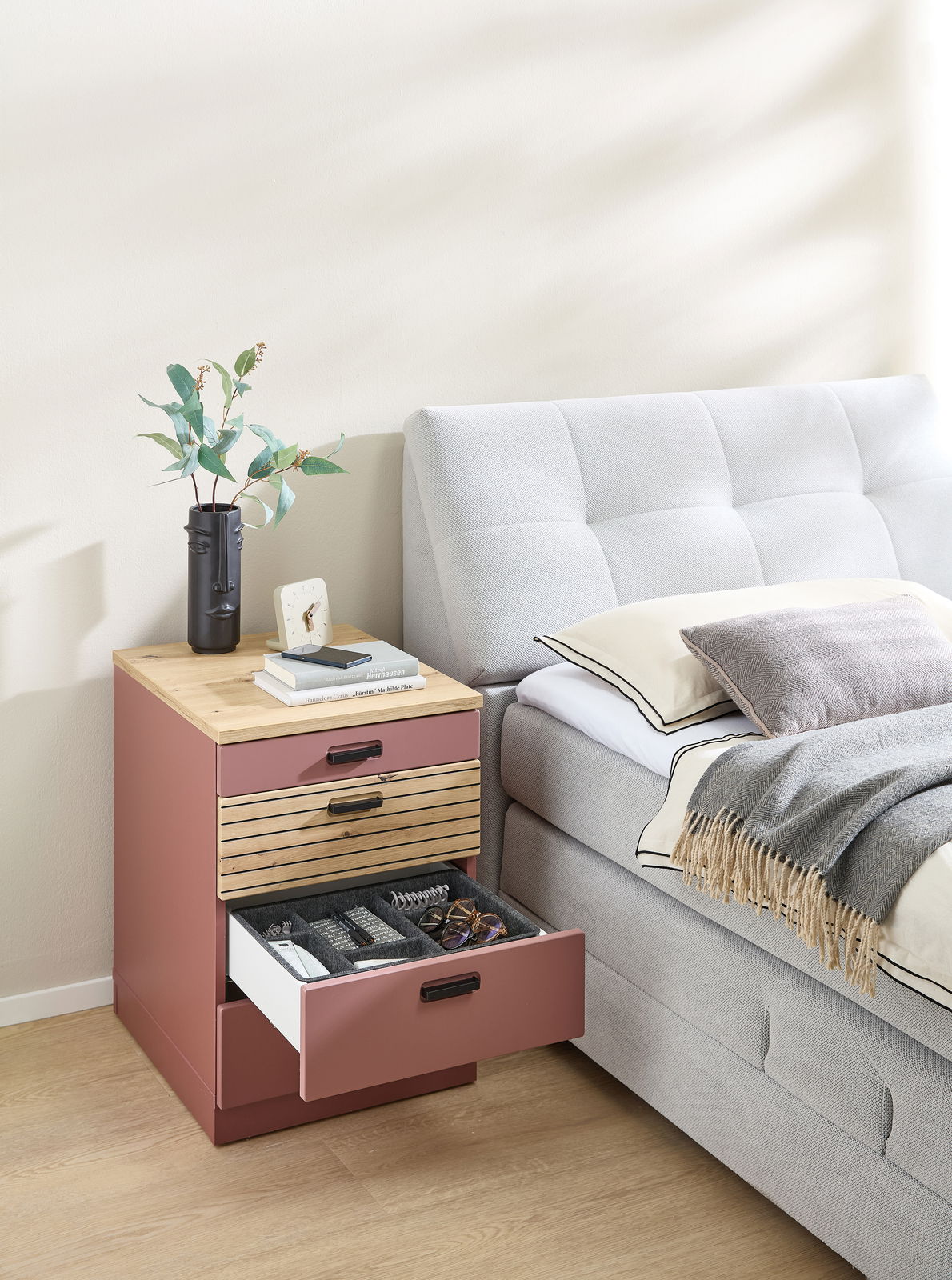 Rollcontainer in einem modernen Schlafzimmer, seitliche Perspektive. Der Container hat eine Holzoberfläche und drei Schubladen, von denen eine geöffnet ist und verschiedene Gegenstände zeigt.