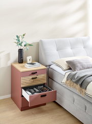 Rollcontainer in einem modernen Schlafzimmer, seitliche Perspektive. Der Container hat eine Holzoberfläche und drei Schubladen, von denen eine geöffnet ist und verschiedene Gegenstände zeigt.