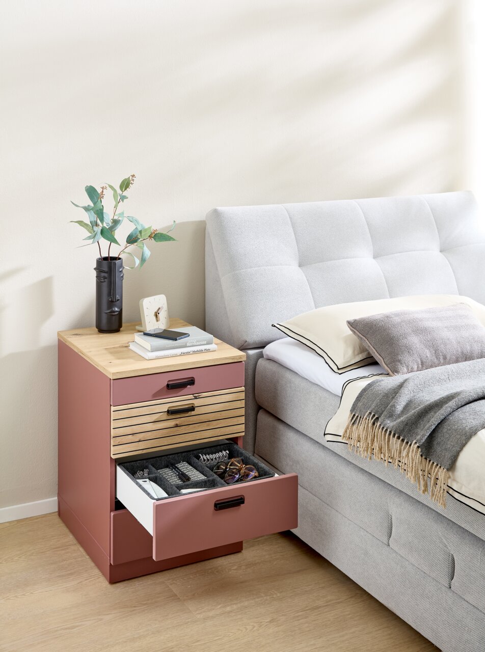 Rollcontainer in einem modernen Schlafzimmer, seitliche Perspektive. Der Container hat eine Holzoberfläche und drei Schubladen, von denen eine geöffnet ist und verschiedene Gegenstände zeigt.