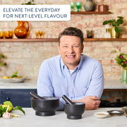 Zwei schwarze Mörser und Stößel auf einer Küchenarbeitsplatte, frontal fotografiert, mit frischen Zutaten und einem lächelnden Mann im Hintergrund.