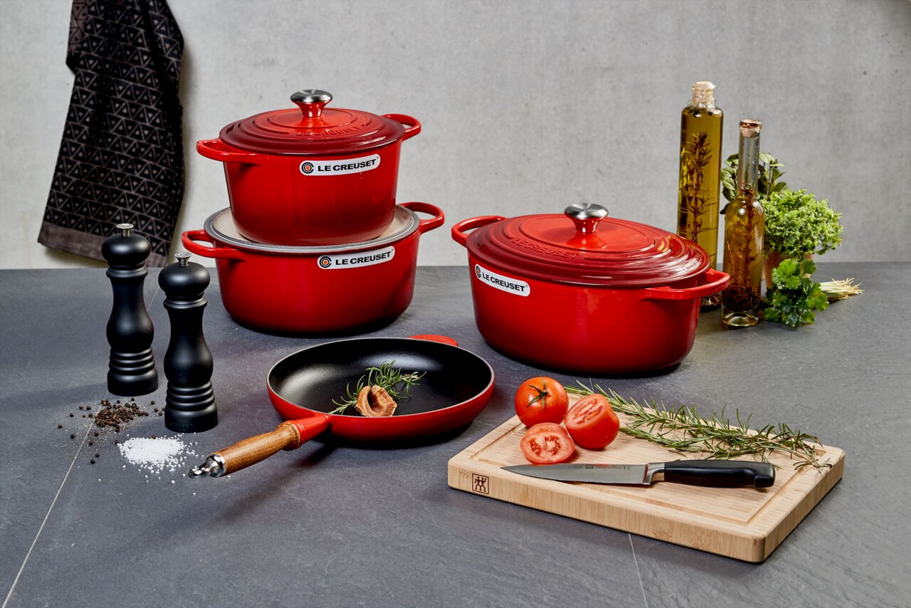 LE CREUSET Bräter kirschrot 33 cm SIGNATURE Küchenszene mit roten Le Creuset Brätern, schwarzer Salzmühle und Pfeffermühle, sowie einer Bratpfanne mit Holzgriff, von vorne betrachtet.