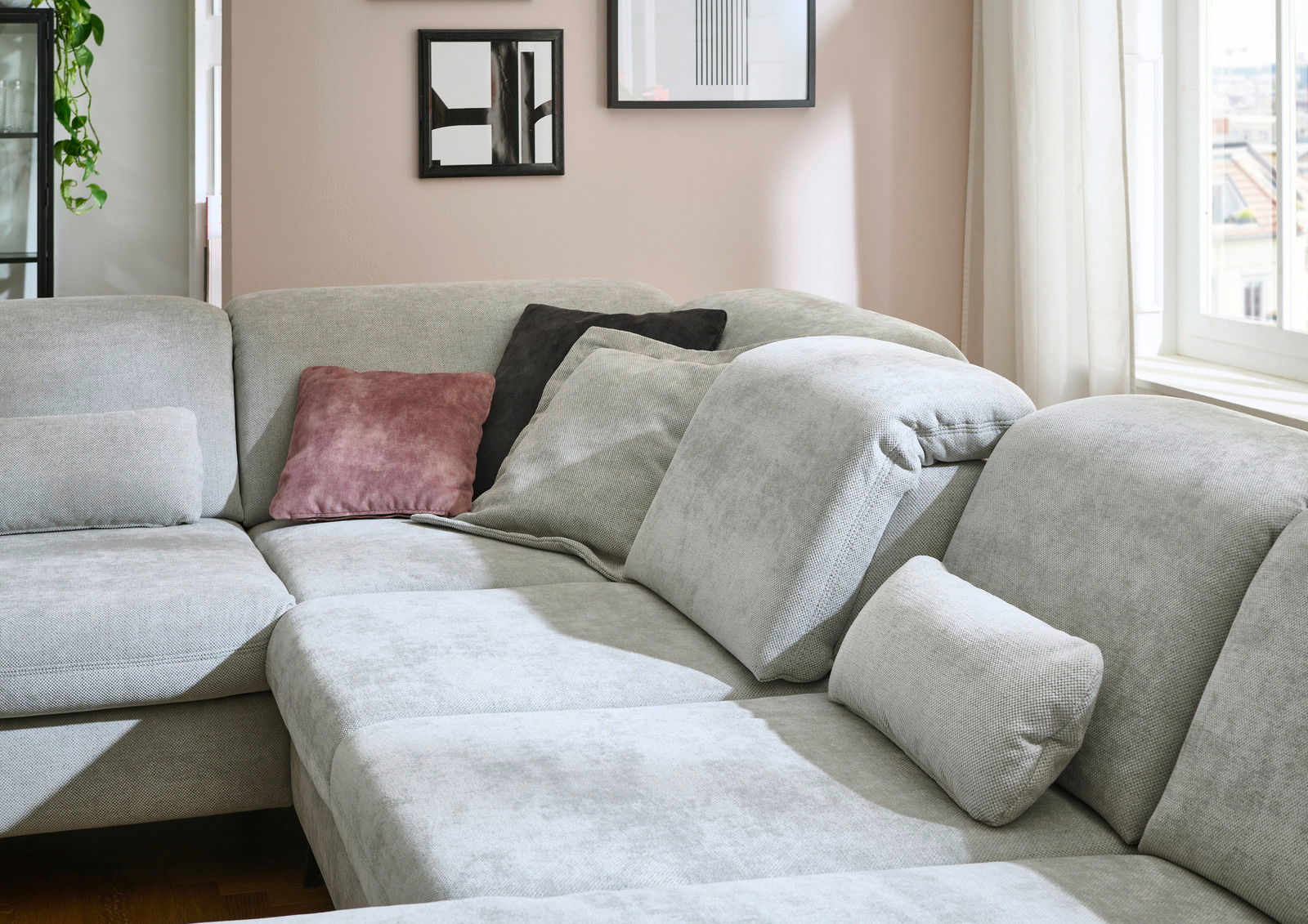 Ecksofa in hellem Grau mit mehreren Kissen in verschiedenen Farben, in einem gemütlichen Wohnzimmer mit Fenstern im Hintergrund. Perspektive von der Seite.