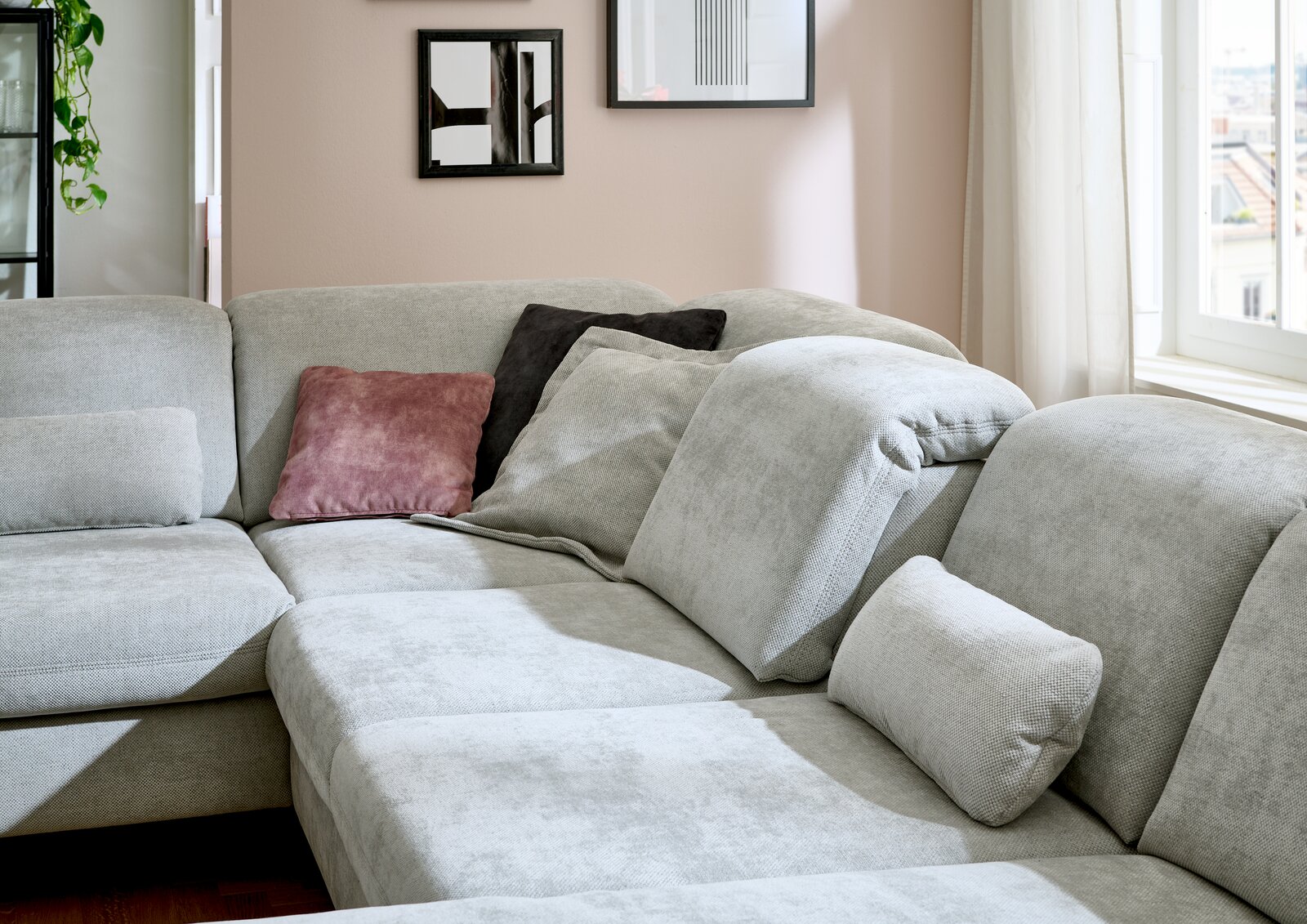 Ecksofa in hellem Grau mit mehreren Kissen in verschiedenen Farben, in einem gemütlichen Wohnzimmer mit Fenstern im Hintergrund. Perspektive von der Seite.