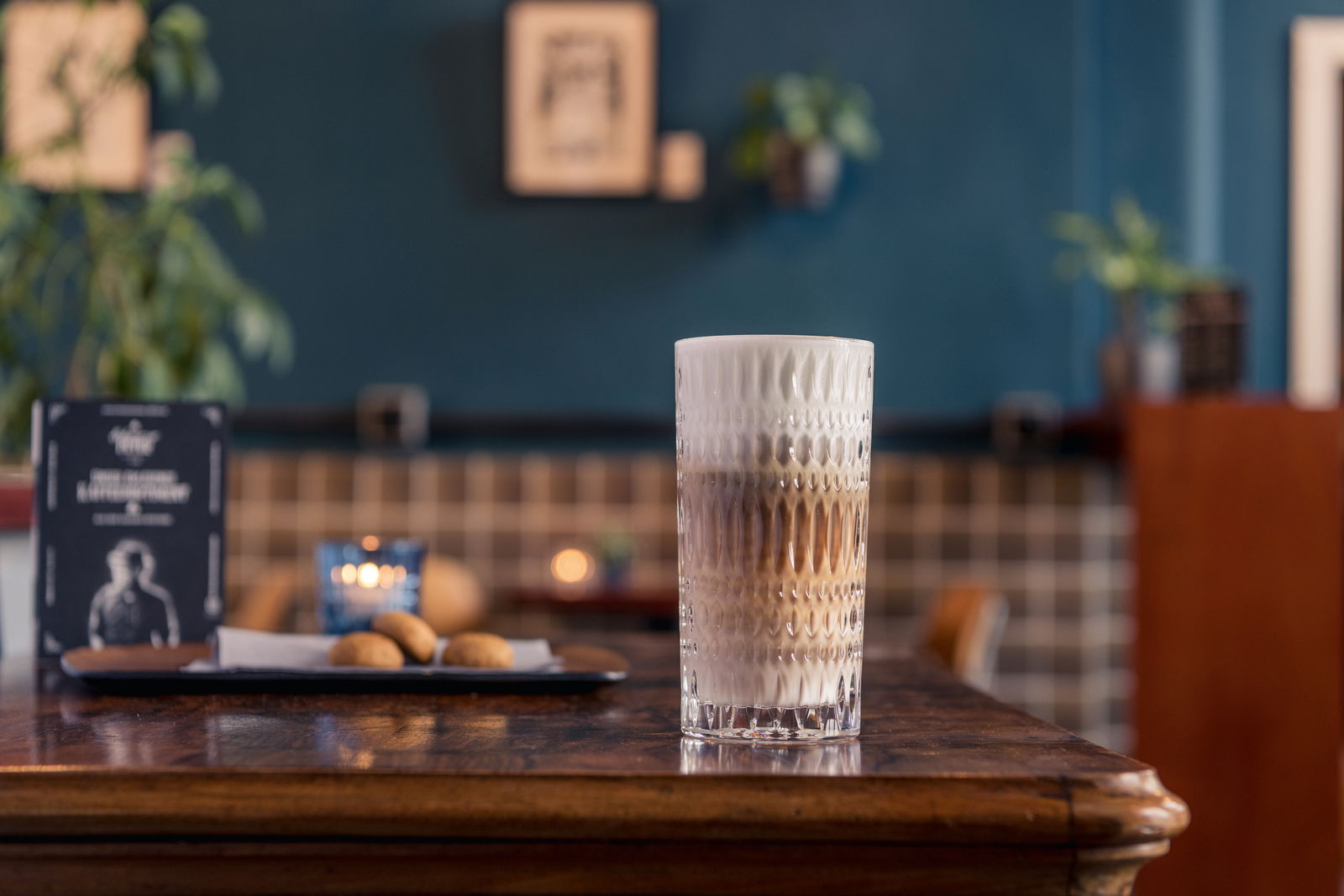 Ein hohes, strukturiertes Glas mit Latte Macchiato auf einem Holztisch in einem gemütlichen Café, seitliche Perspektive.