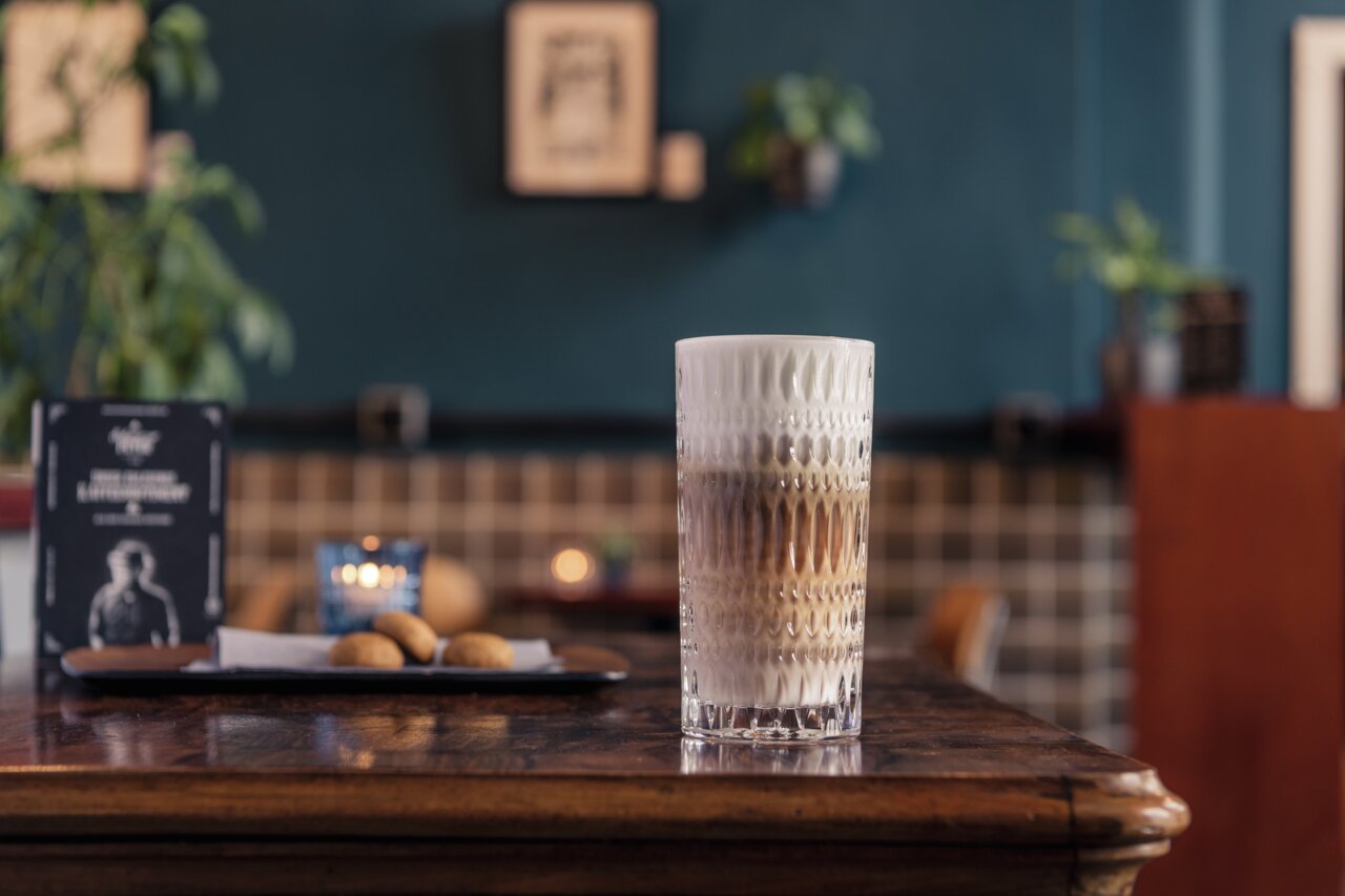 Ein hohes, strukturiertes Glas mit Latte Macchiato auf einem Holztisch in einem gemütlichen Café, seitliche Perspektive.