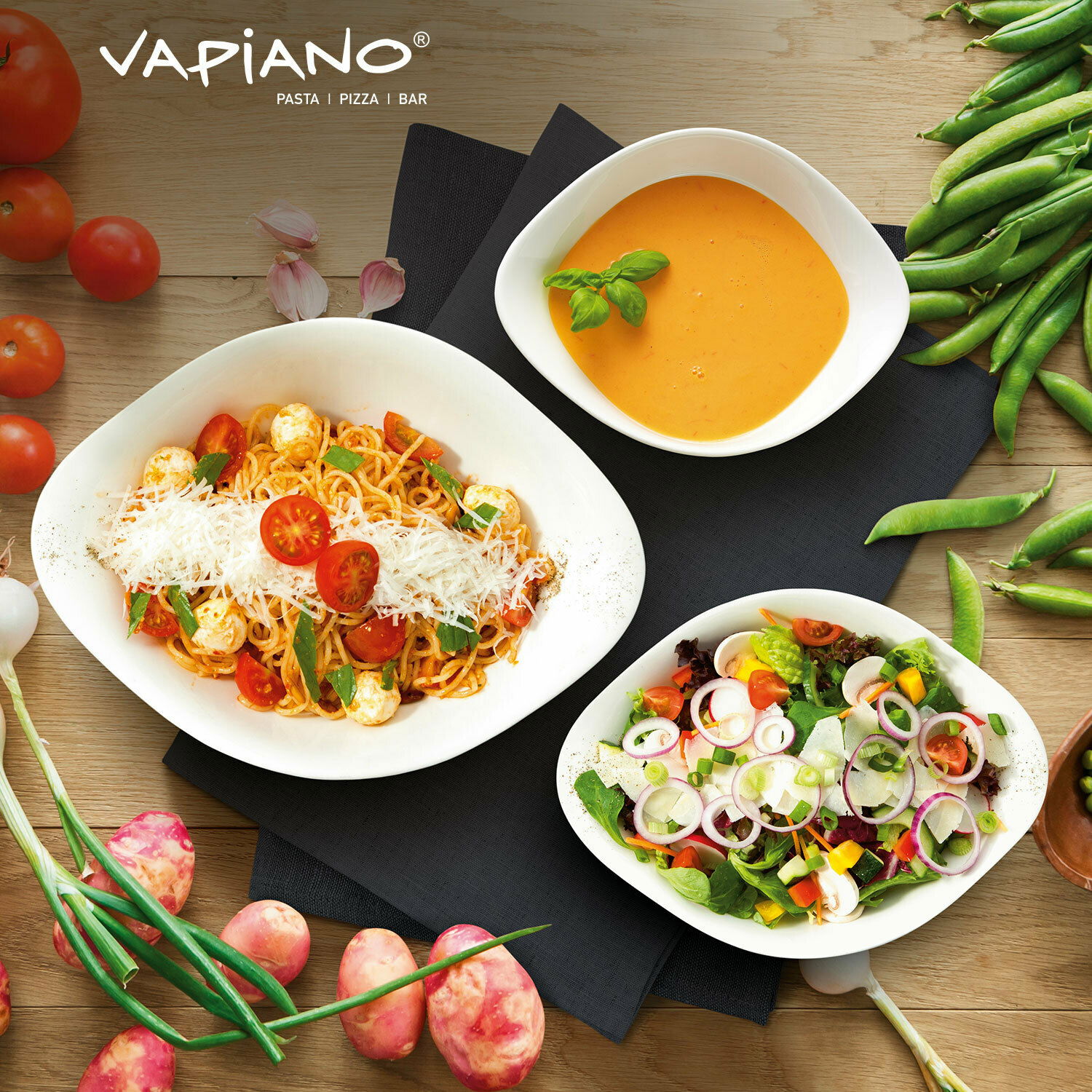 Draufsicht auf ein Vapiano Starter-Set mit drei weißen Schalen, gefüllt mit Spaghetti, Suppe und Salat, umgeben von frischen Zutaten wie Tomaten und Erbsen.