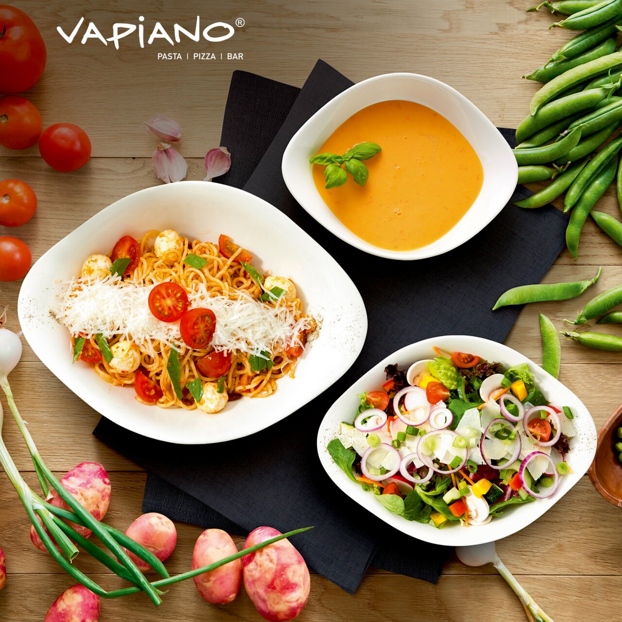 Draufsicht auf ein Vapiano Starter-Set mit drei weißen Schalen, gefüllt mit Spaghetti, Suppe und Salat, umgeben von frischen Zutaten wie Tomaten und Erbsen.