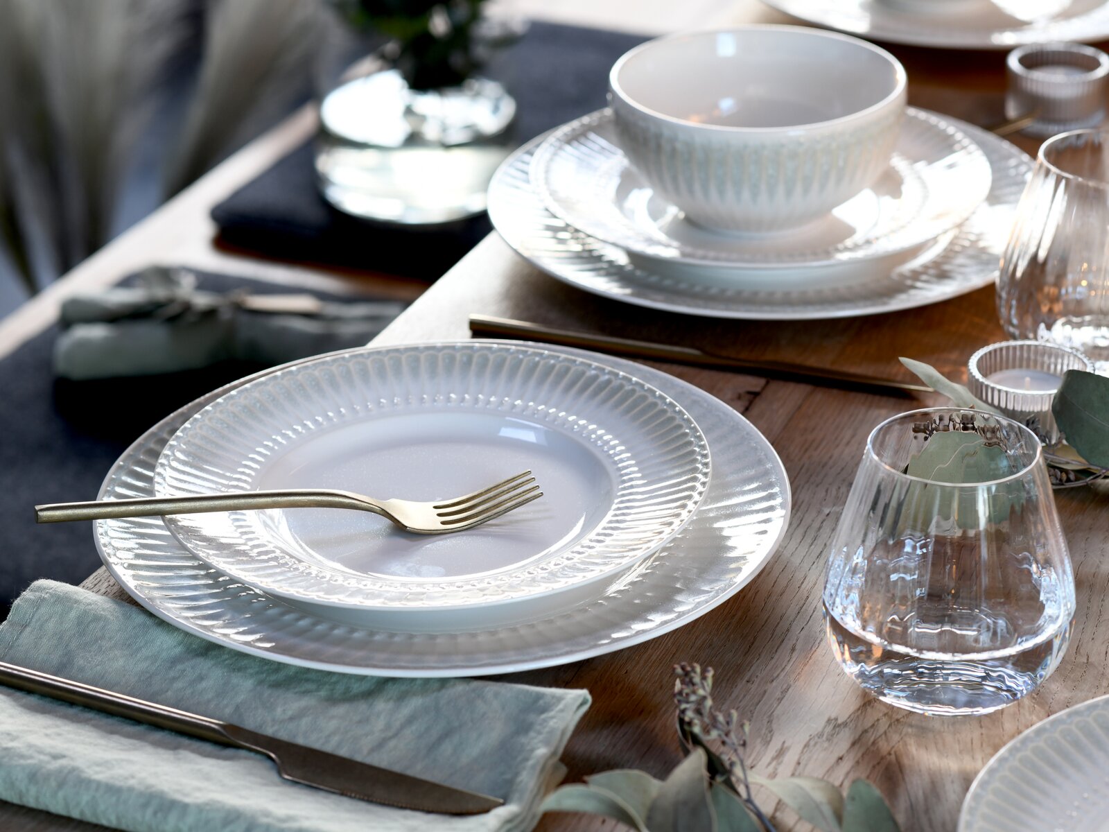 Elegantes weißes Porzellan-Gedeck mit geriffeltem Teller und Schale auf Holztisch, goldfarbenem Besteck und Wasserglas, fotografiert aus schräger Draufsicht.