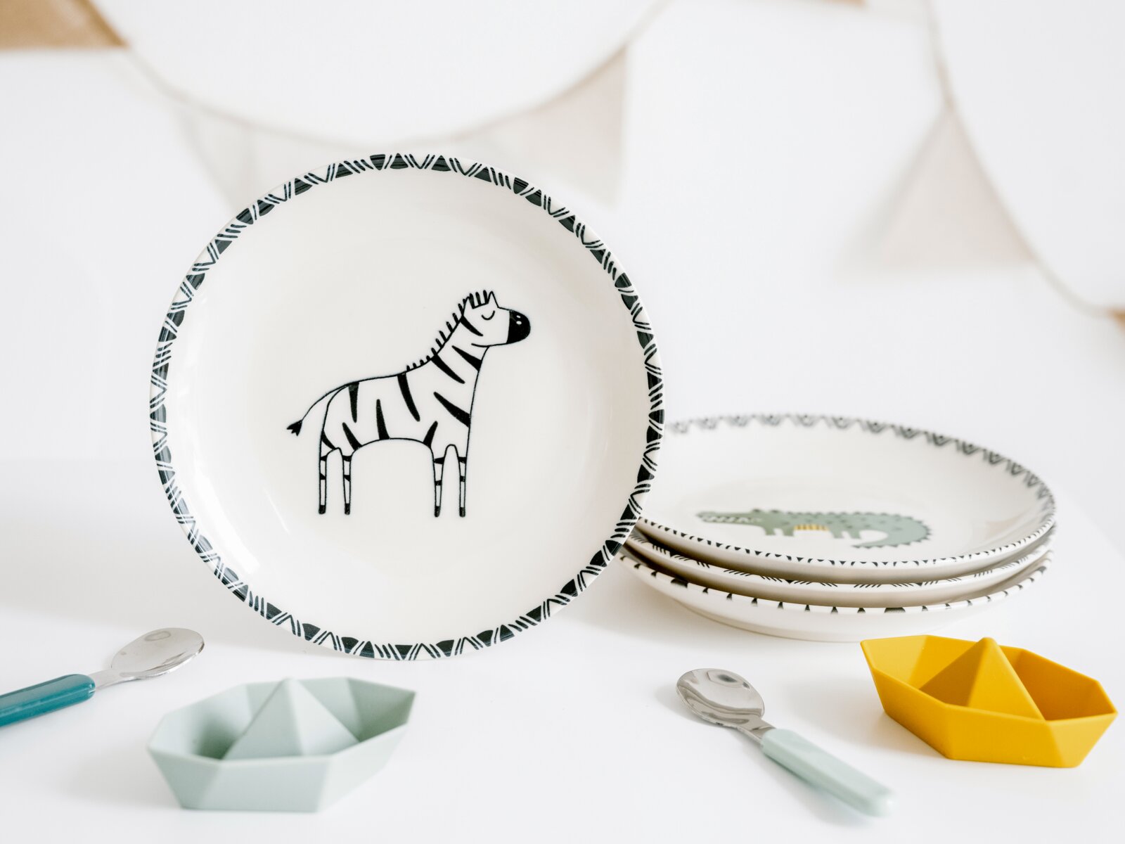 Keramikteller in Weiß mit Zebra-Motiv und gemustertem Rand, frontal aus leicht schräger Perspektive fotografiert; daneben gestapelte Teller mit Krokodil-Motiv sowie Kinderlöffel und kleine Schälchen auf weißem Hintergrund.