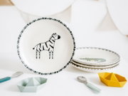 Keramikteller in Weiß mit Zebra-Motiv und gemustertem Rand, frontal aus leicht schräger Perspektive fotografiert; daneben gestapelte Teller mit Krokodil-Motiv sowie Kinderlöffel und kleine Schälchen auf weißem Hintergrund.