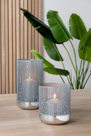 Zwei moderne Windlichter aus Metall mit geometrischem Muster, auf einem Holztisch stehend, im Hintergrund grüne Pflanzen. Perspektive von vorne.