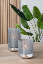 Zwei moderne Windlichter aus Metall mit geometrischem Muster, auf einem Holztisch stehend, im Hintergrund grüne Pflanzen. Perspektive von vorne.