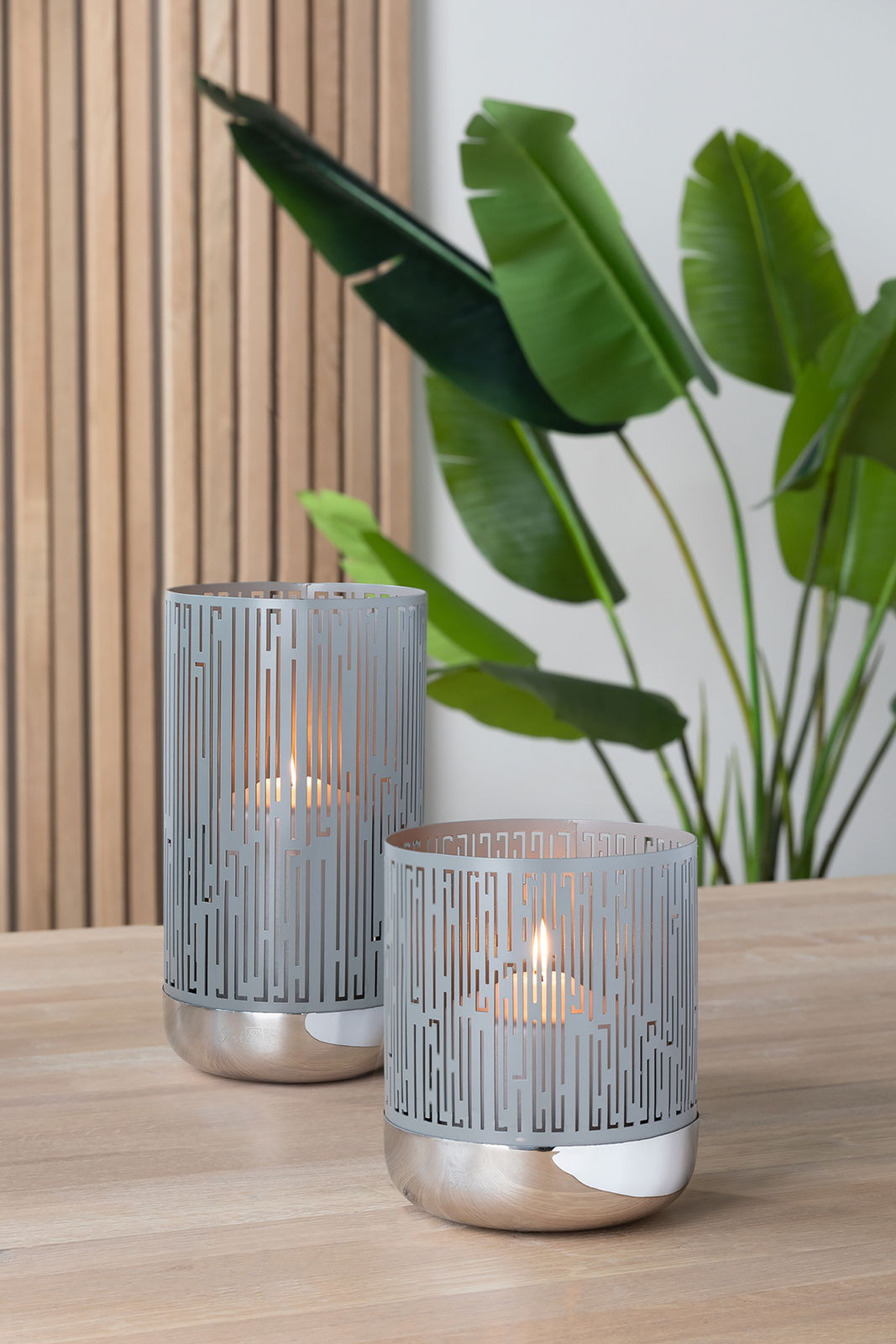 Fink Windlicht BRIANO Zwei moderne Windlichter aus Metall mit geometrischem Muster, auf einem Holztisch stehend, im Hintergrund grüne Pflanzen. Perspektive von vorne.