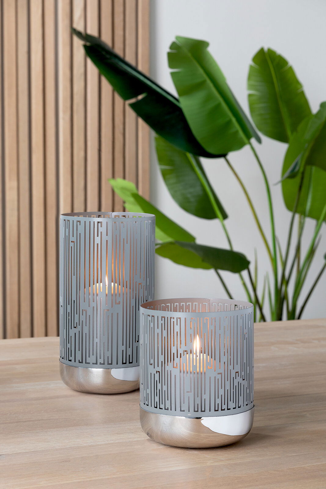 Zwei moderne Windlichter aus Metall mit geometrischem Muster, auf einem Holztisch stehend, im Hintergrund grüne Pflanzen. Perspektive von vorne.