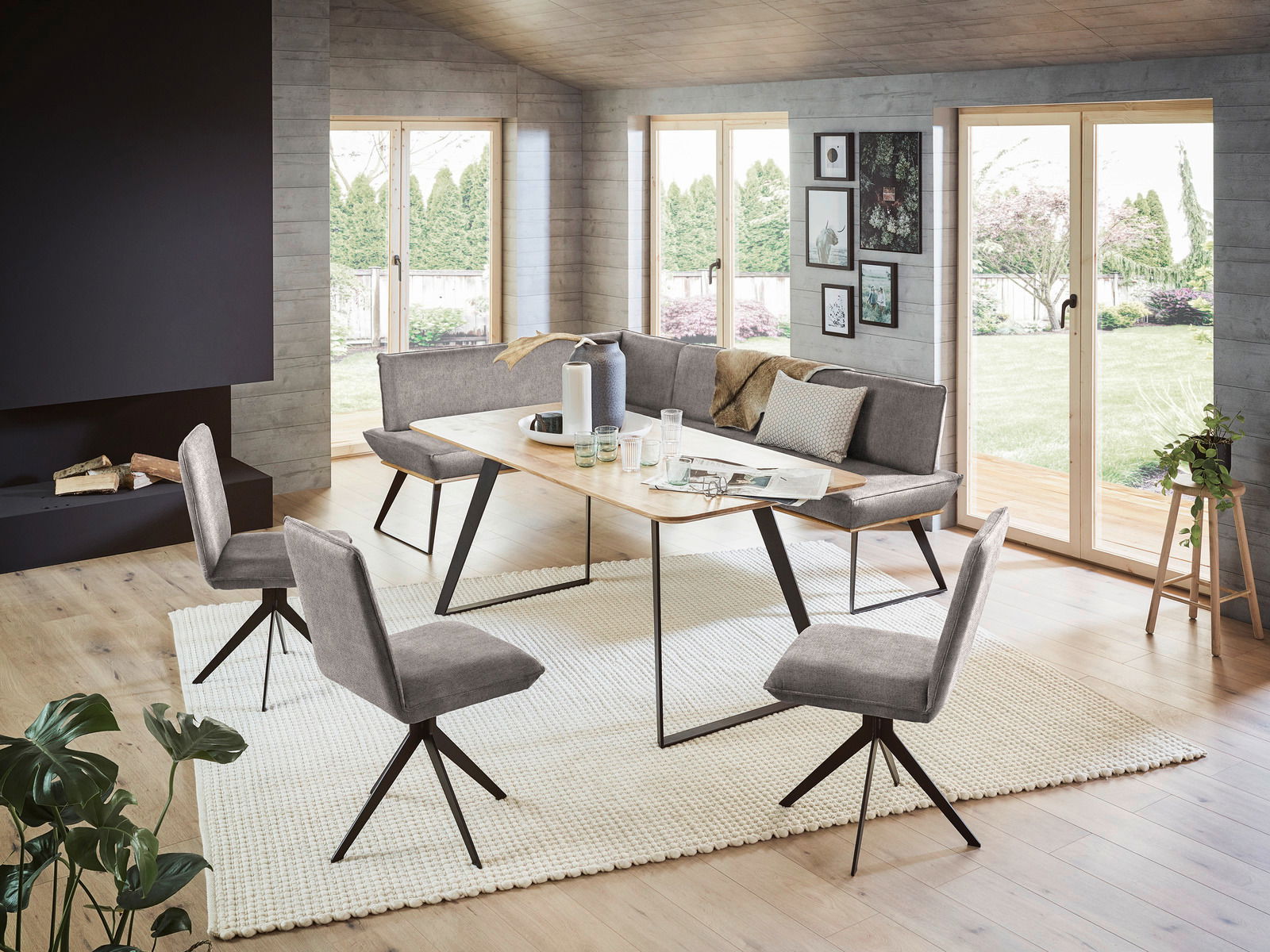 Voleo Eckbank MALIBU Moderne Esszimmer-Eckbankgruppe mit grauen Polstern und einem Holztisch, umgeben von großen Fenstern mit Blick auf den Garten. Perspektive von schräg vorne.