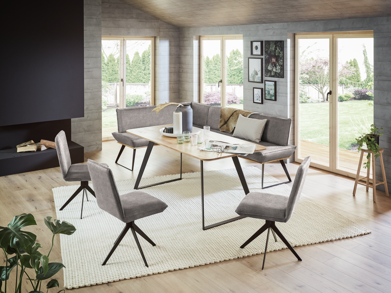 Moderne Esszimmer-Eckbankgruppe mit grauen Polstern und einem Holztisch, umgeben von großen Fenstern mit Blick auf den Garten. Perspektive von schräg vorne.