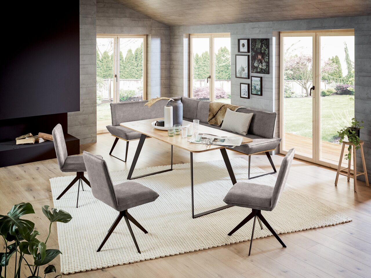 Voleo Eckbank MALIBU Moderne Esszimmer-Eckbankgruppe mit grauen Polstern und einem Holztisch, umgeben von großen Fenstern mit Blick auf den Garten. Perspektive von schräg vorne.