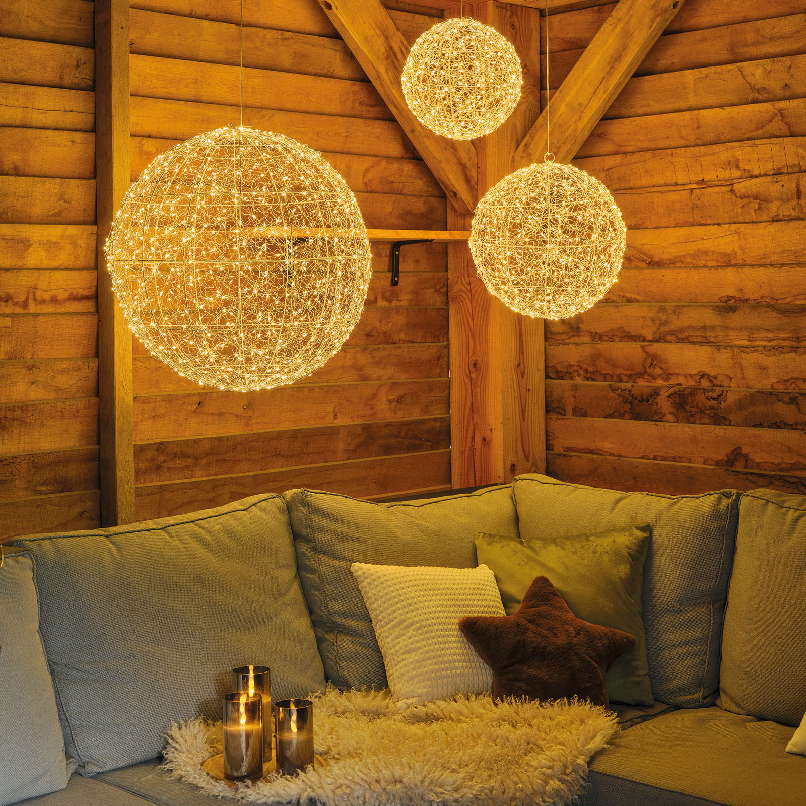 Gemütliche Wohnzimmeratmosphäre mit hängenden LED-Kugeln in verschiedenen Größen, die warmes Licht spenden, im Vordergrund ein Sofa mit Kissen und Kerzen auf einem Tisch, seitliche Perspektive.