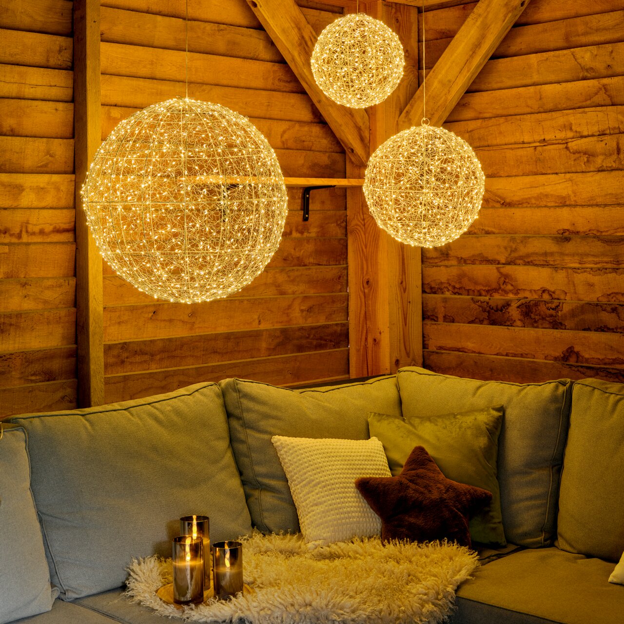 Gemütliche Wohnzimmeratmosphäre mit hängenden LED-Kugeln in verschiedenen Größen, die warmes Licht spenden, im Vordergrund ein Sofa mit Kissen und Kerzen auf einem Tisch, seitliche Perspektive.