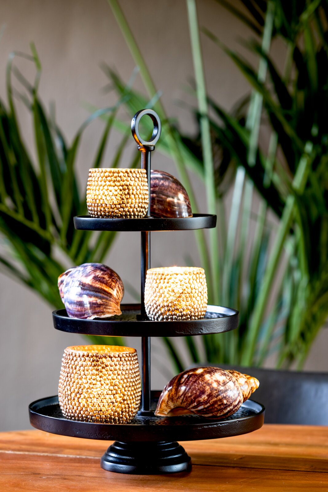 Schwarze dreistöckige Metall-Etagere mit Ringgriff, dekoriert mit goldfarbenen Windlichtern und großen Muscheln, in leichter Frontalansicht von oben