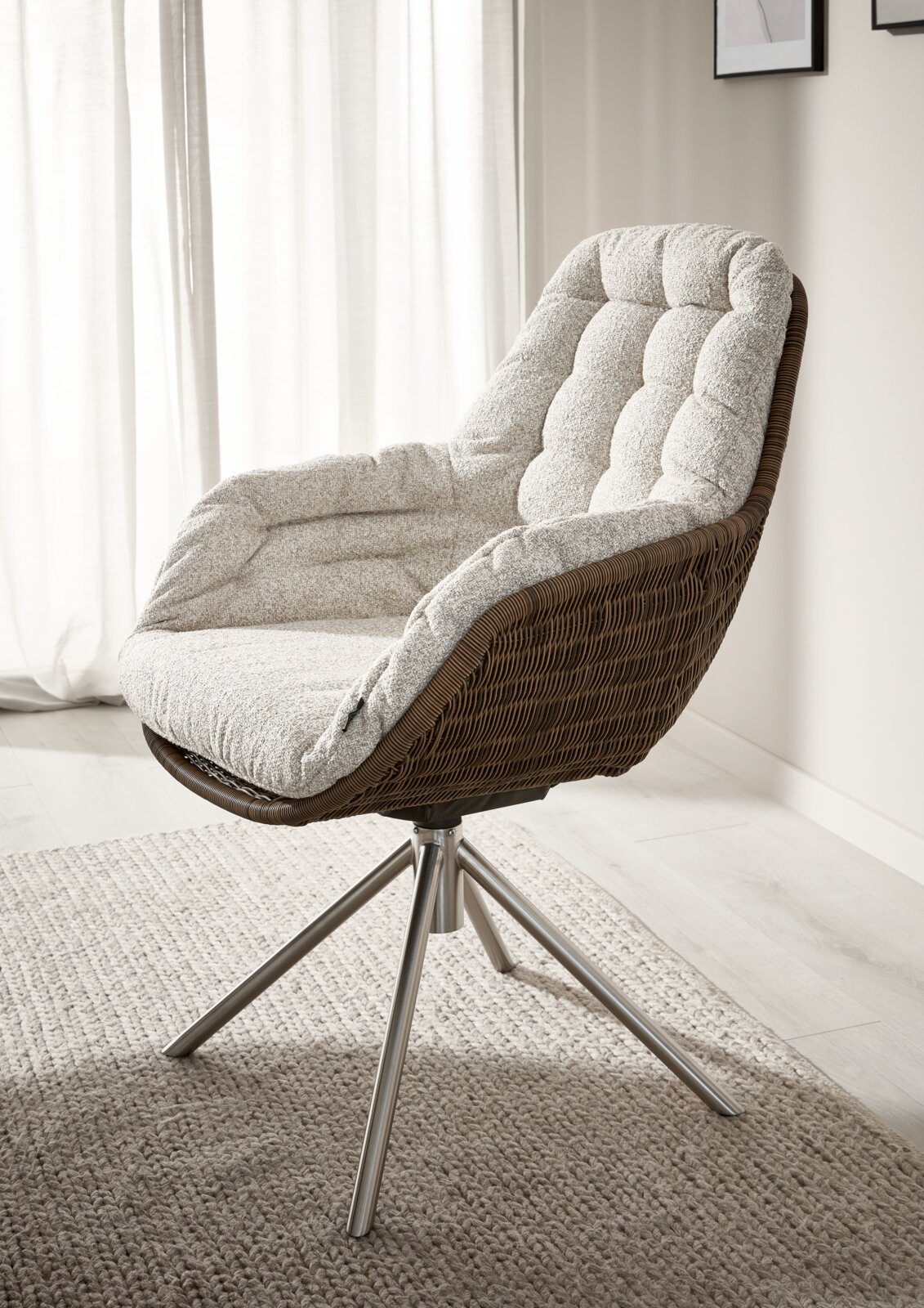 Johann Jakob Armlehnstuhl CURBI Bequemer Lounge-Drehsessel mit braunem Rattan-/Korbflecht-Gestell, hellgrau gepolstertem Sitzkissen und silbernem Metallfuß, im Wohnzimmer vor hellen Vorhängen – seitliche 3/4-Ansicht von vorne rechts.