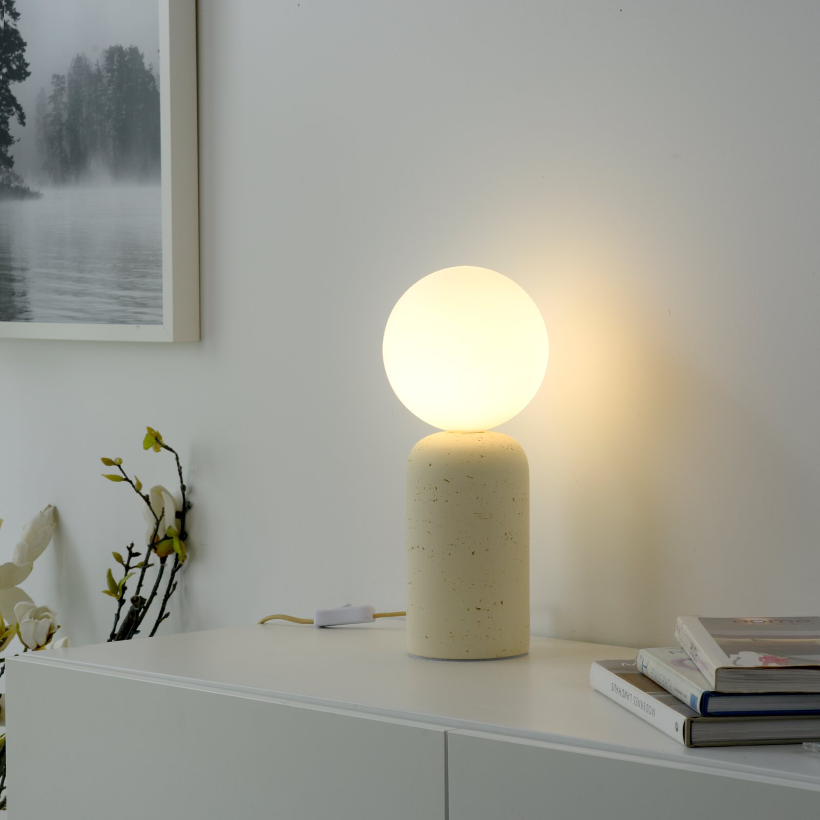Leuchtende Tischlampe mit kugelförmigem, opalem Schirm auf zylindrischem, hell gesprenkeltem Sockel auf weißem Sideboard, frontale Perspektive mit leichter Seitenansicht.