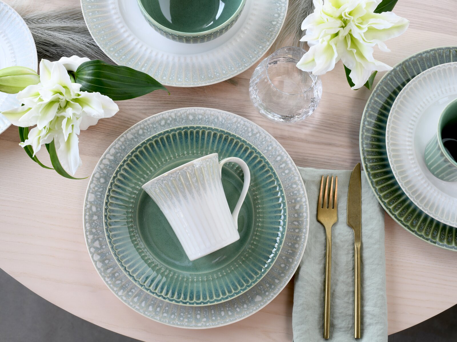 Draufsicht auf ein stilvolles Tischgedeck mit grün-weiß glasierten Keramiktellern, einer weißen geriffelten Tasse auf dem Teller, goldfarbenem Besteck auf einer mintgrünen Serviette, Wasserglas und weißen Blumen auf hellem Holztisch.