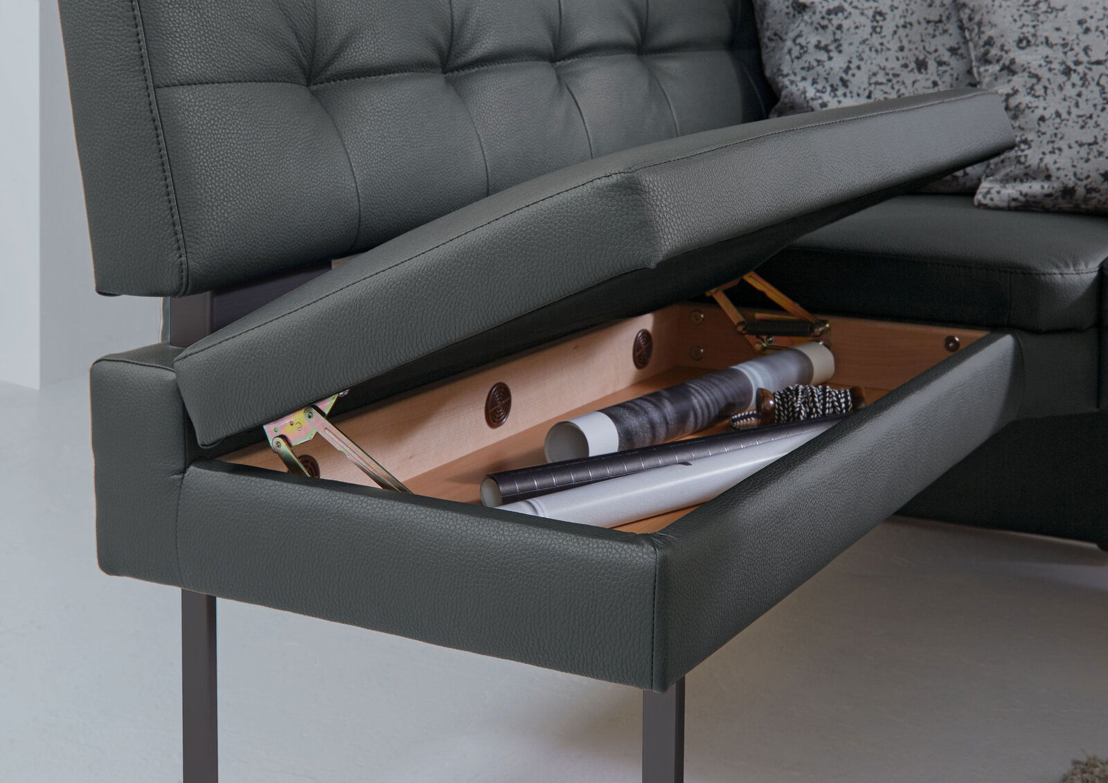 Nahaufnahme einer Eckbank mit aufklappbarem Sitzpolster, das Stauraum für verschiedene Gegenstände bietet. Perspektive von der Seite.